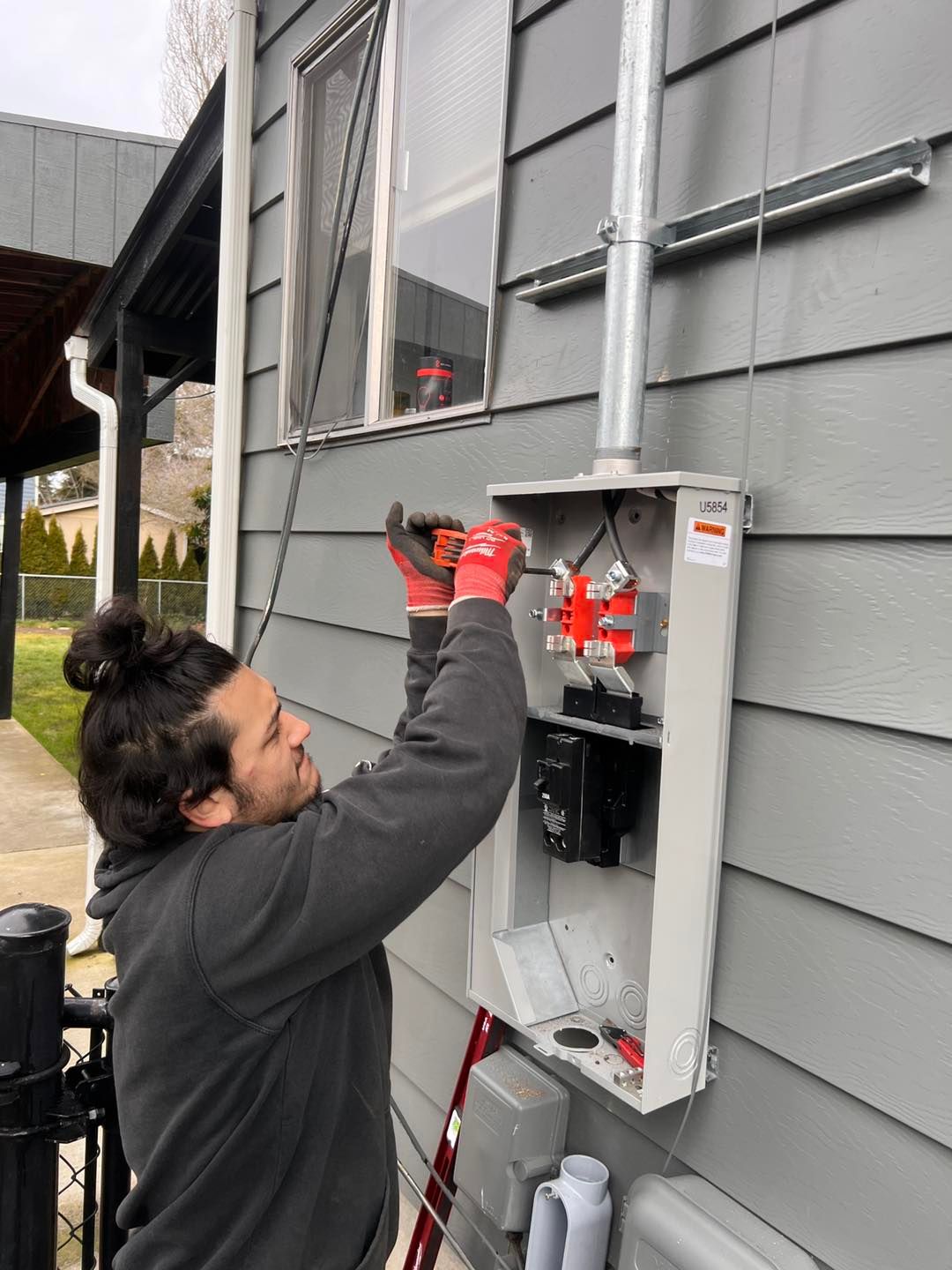 Person in a grey sweatshirt working on an electrical box outside a building, using a screwdriver.