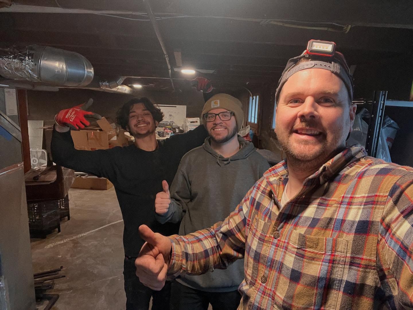 Three people in a basement, smiling and giving thumbs up. One has a headlamp and plaid shirt.