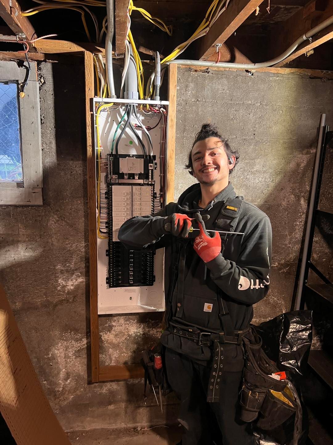 Electrician smiles, pointing at an electrical panel in a basement. Wires exposed; wearing work clothes and tool belt.