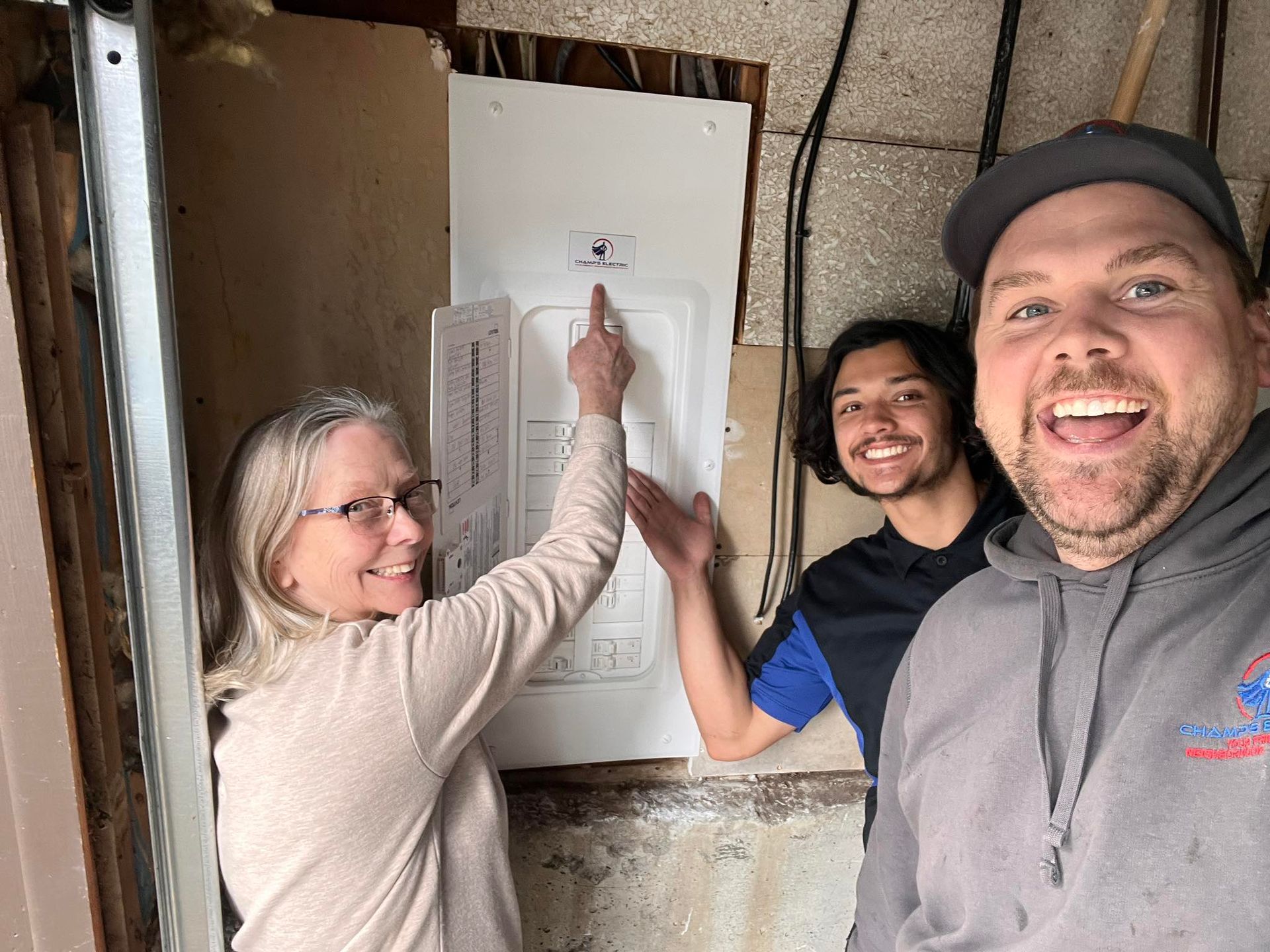 Three people smiling, pointing at an electrical panel on a wall.