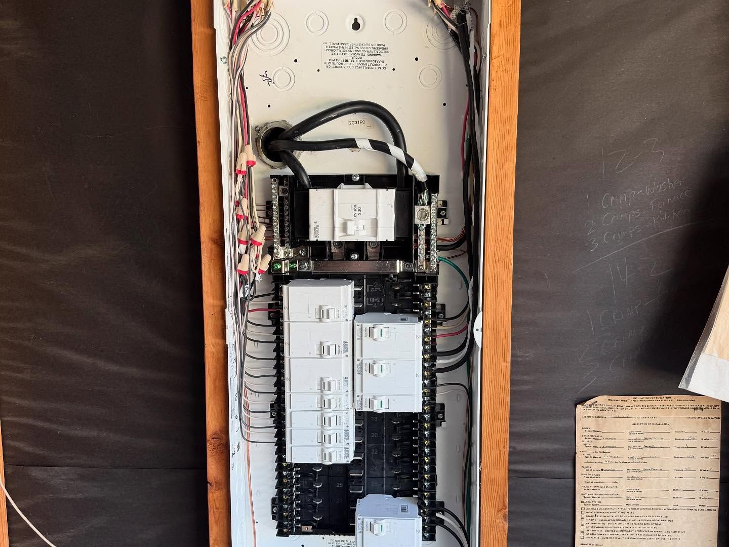 Electrical panel with numerous breakers and wiring inside a wooden frame.