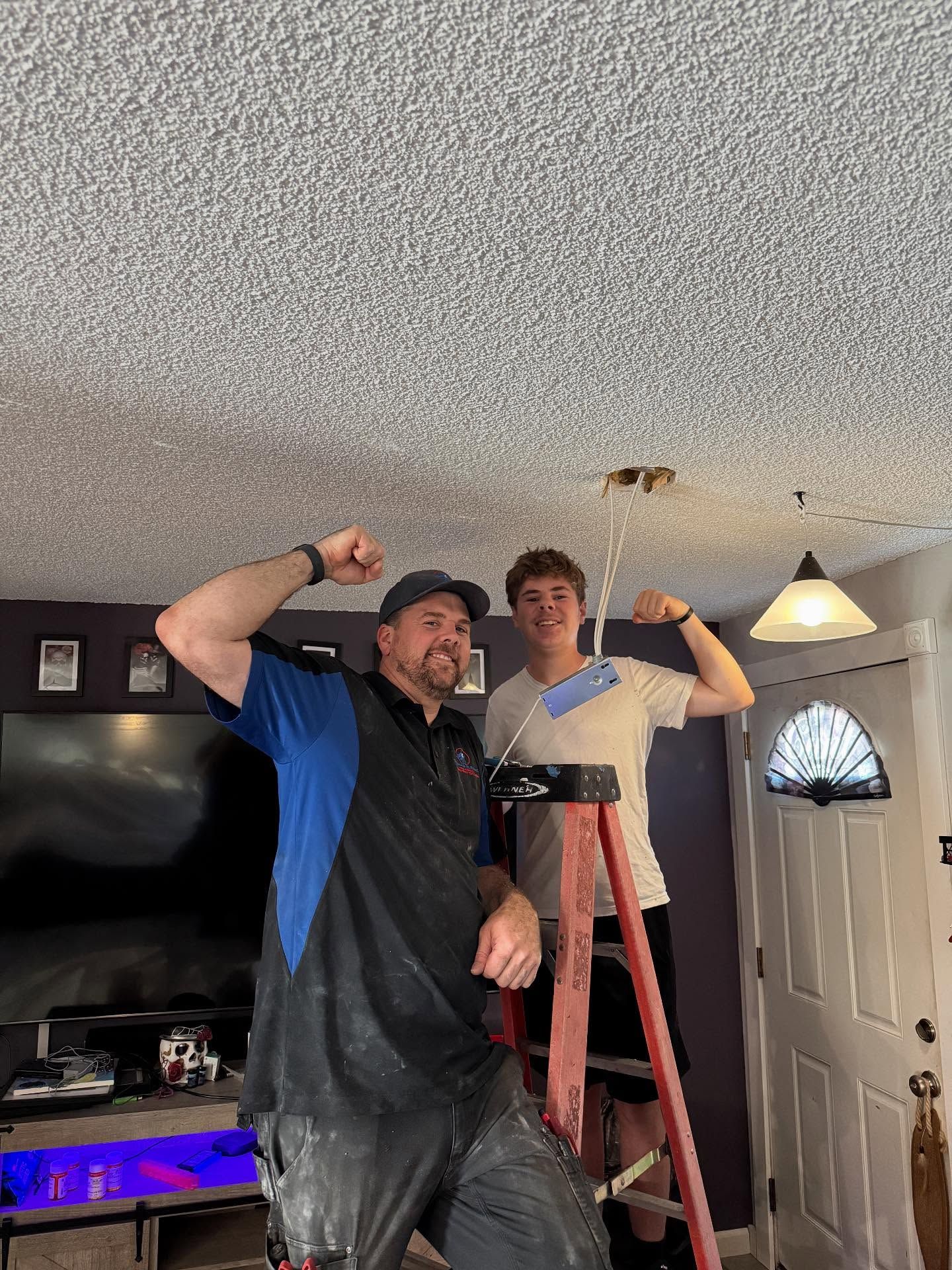 Two people flex arms near a ceiling, one on a ladder. A light fixture hangs above.