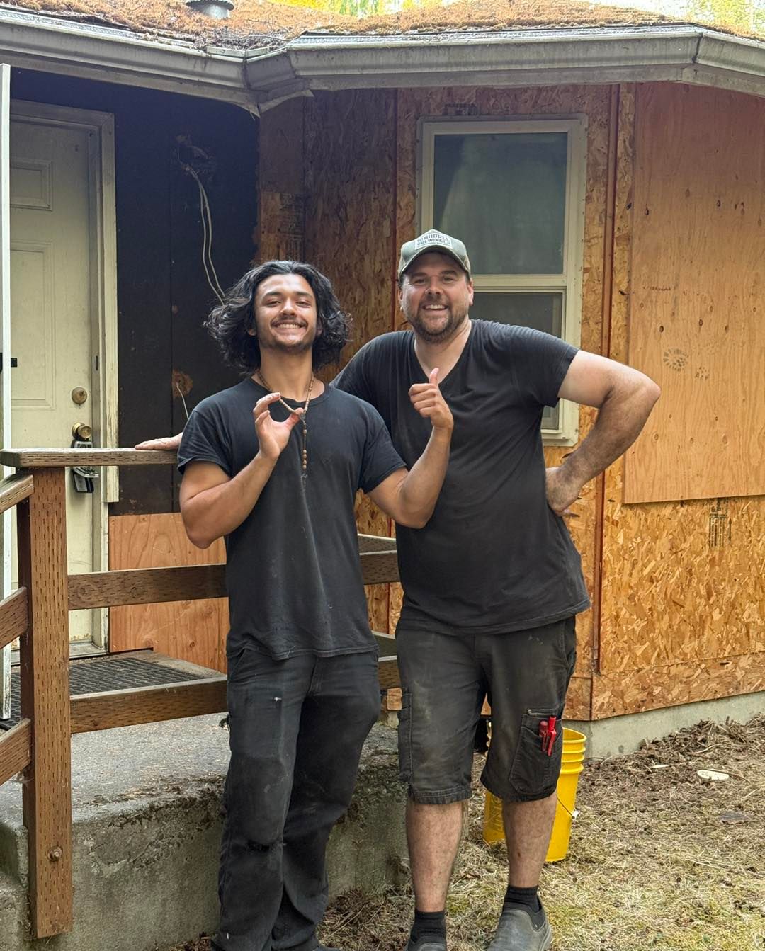 Two men pose in front of a house. One gives the 