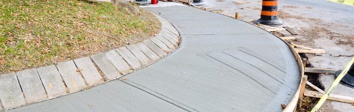 Curved concrete sidewalk under construction beside a grassy verge and orange traffic barrels