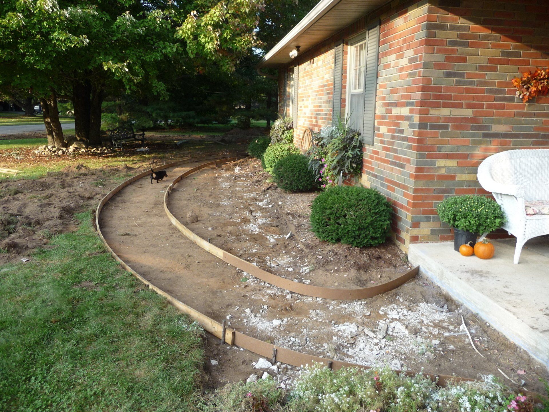 Curved concrete walkway beside a brick house with shrubs and a white chair in the yard