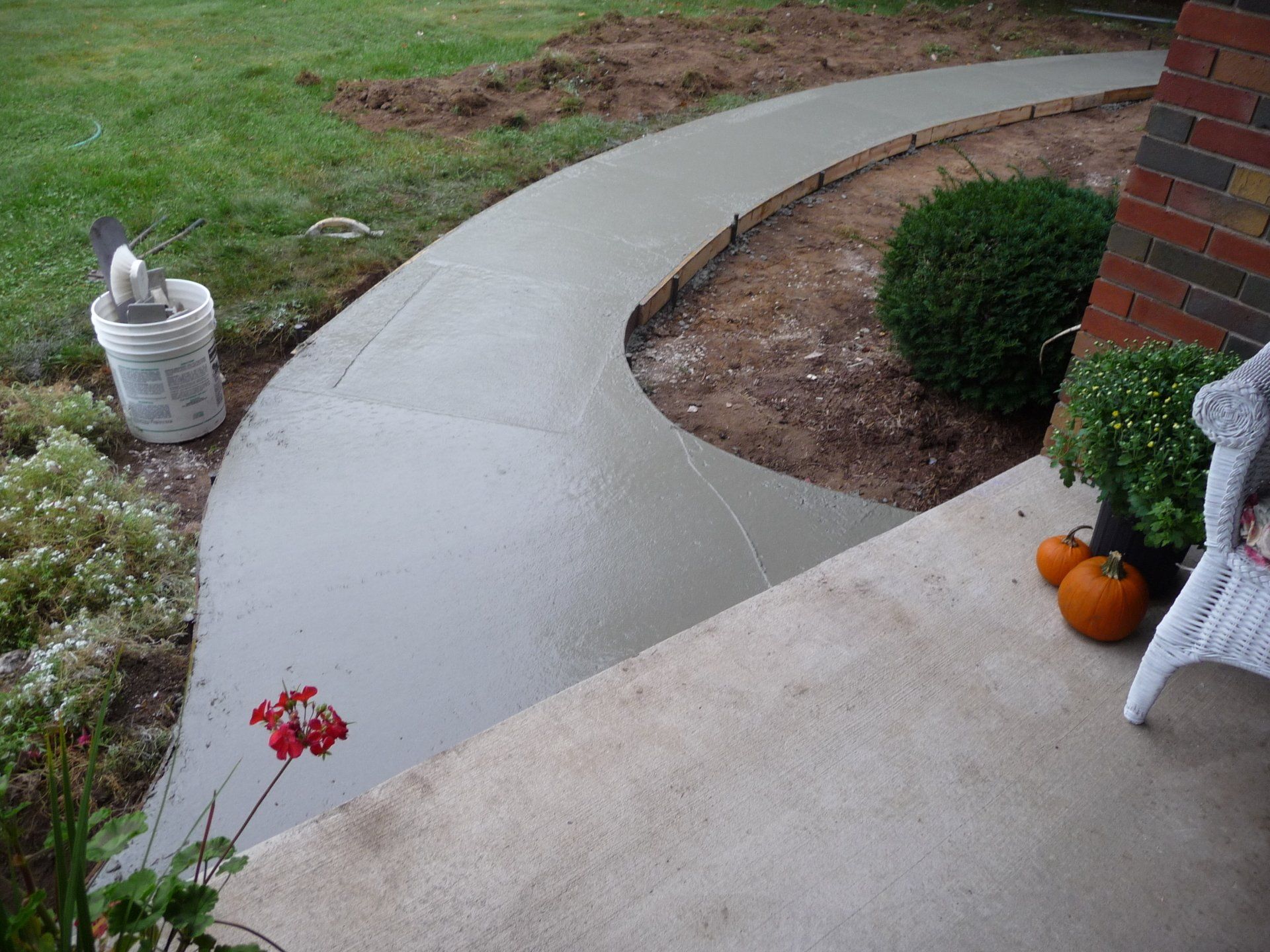 Curved concrete walkway beside a brick house, with a small pumpkin and flower pot near the steps.