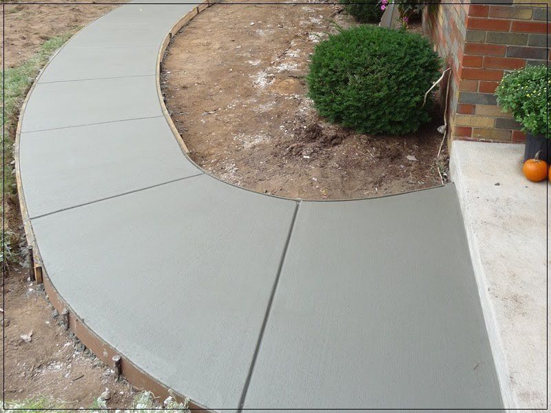 Curved light-gray concrete walkway around a dirt bed and shrub beside a brick house entrance
