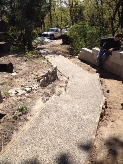 Concrete path through a wooded area with a parked white vehicle and a person sitting on a low wall.