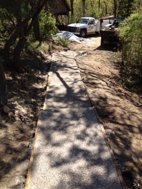Narrow concrete path through a wooded area, leading to parked trucks and a worksite ahead