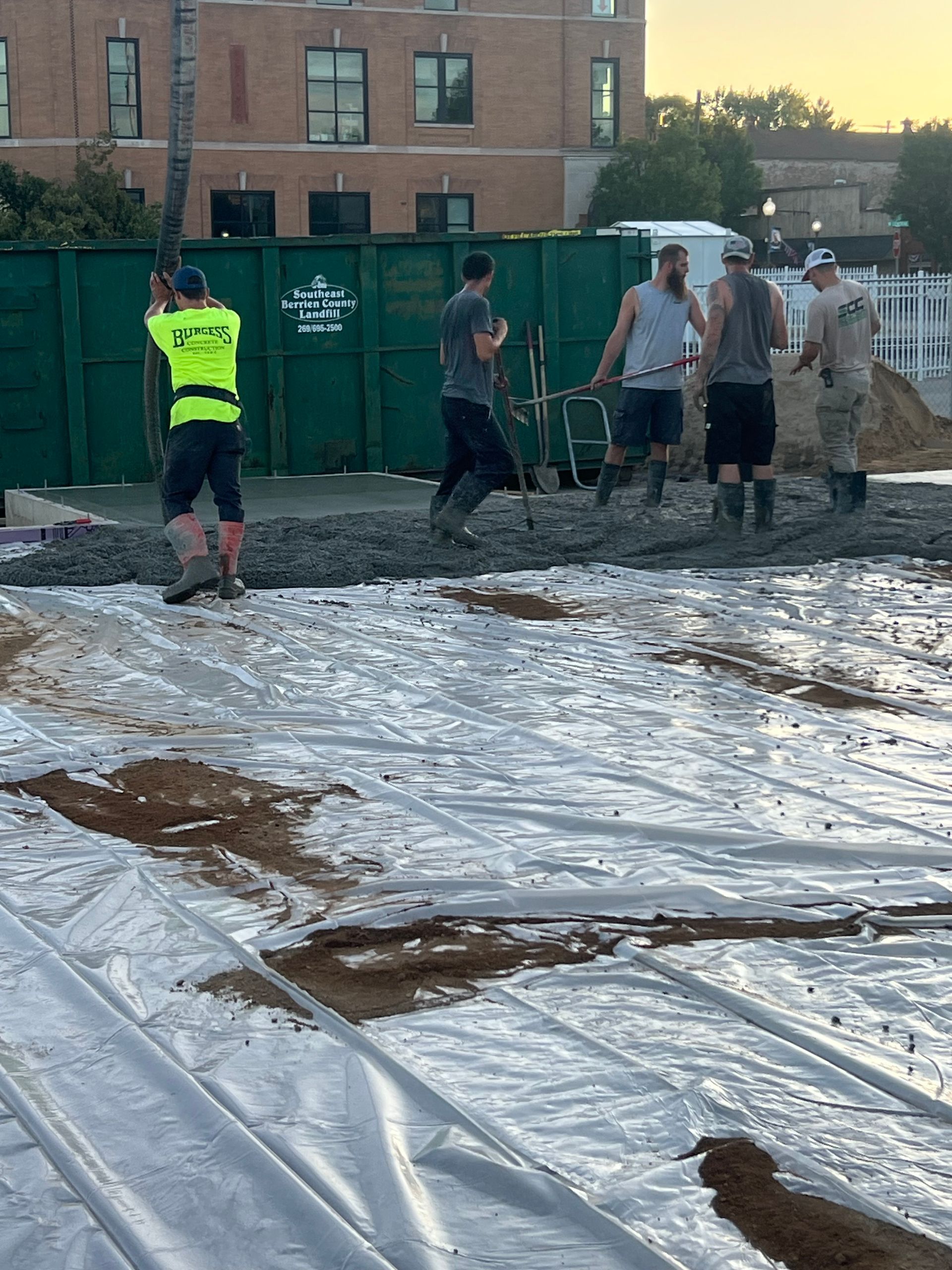 Workers standing on a tarp-covered ground beside green dumpsters at a construction site at dusk