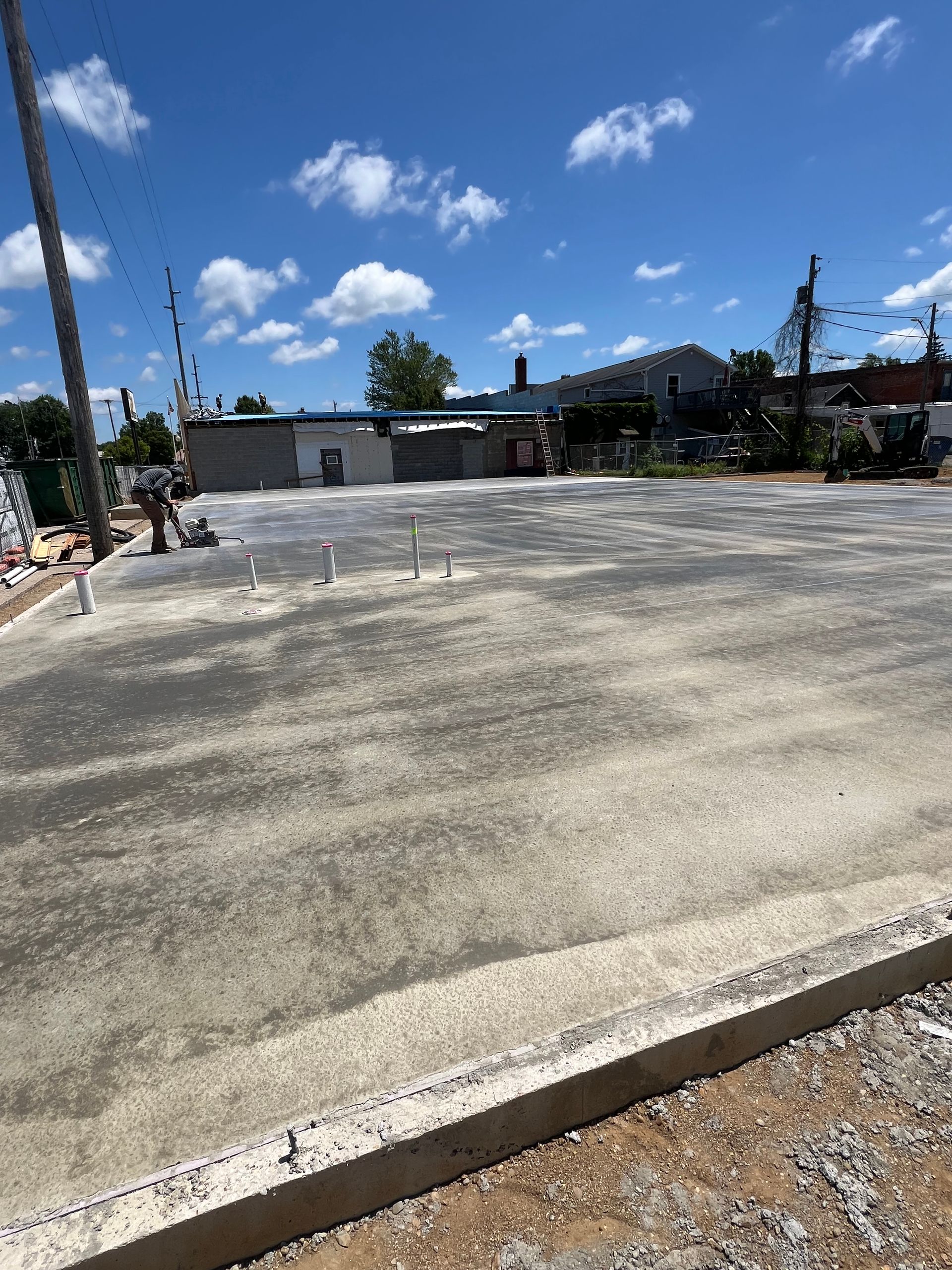 Empty paved lot under a bright blue sky with clouds, bordered by a curb and small buildings.