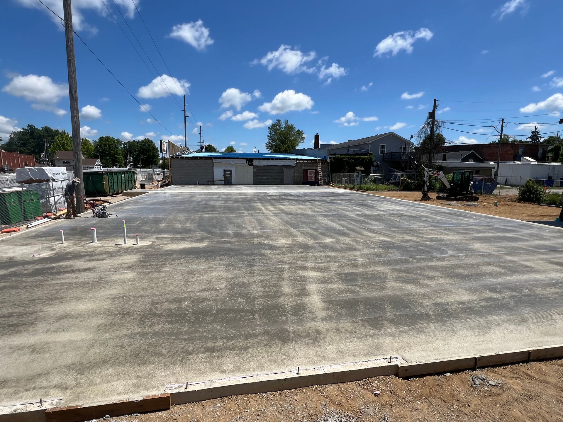 Empty concrete lot under a bright blue sky with clouds, bordered by low buildings and trees