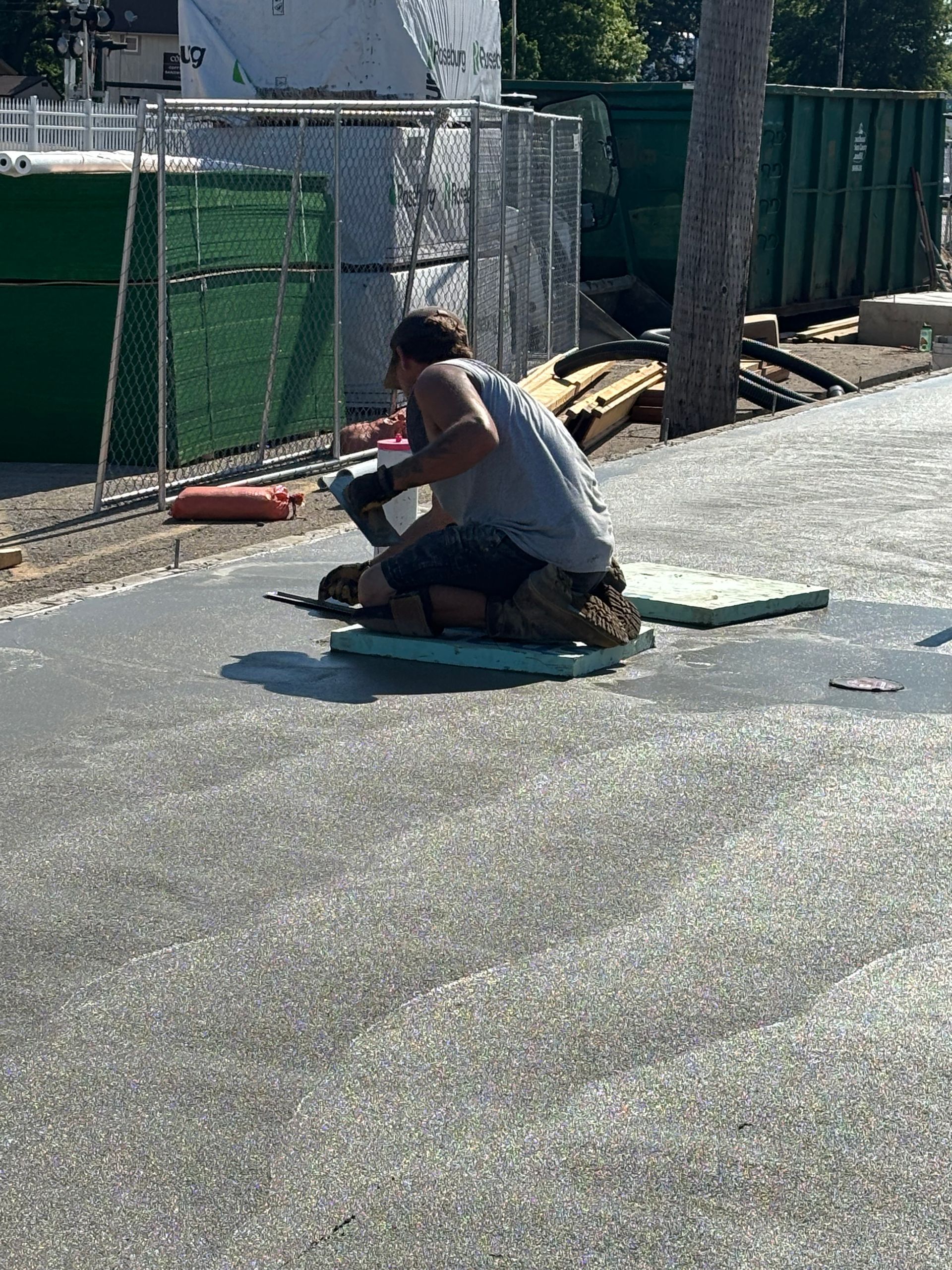 Person crouching on a rooftop, working beside a green fence in bright sunlight.