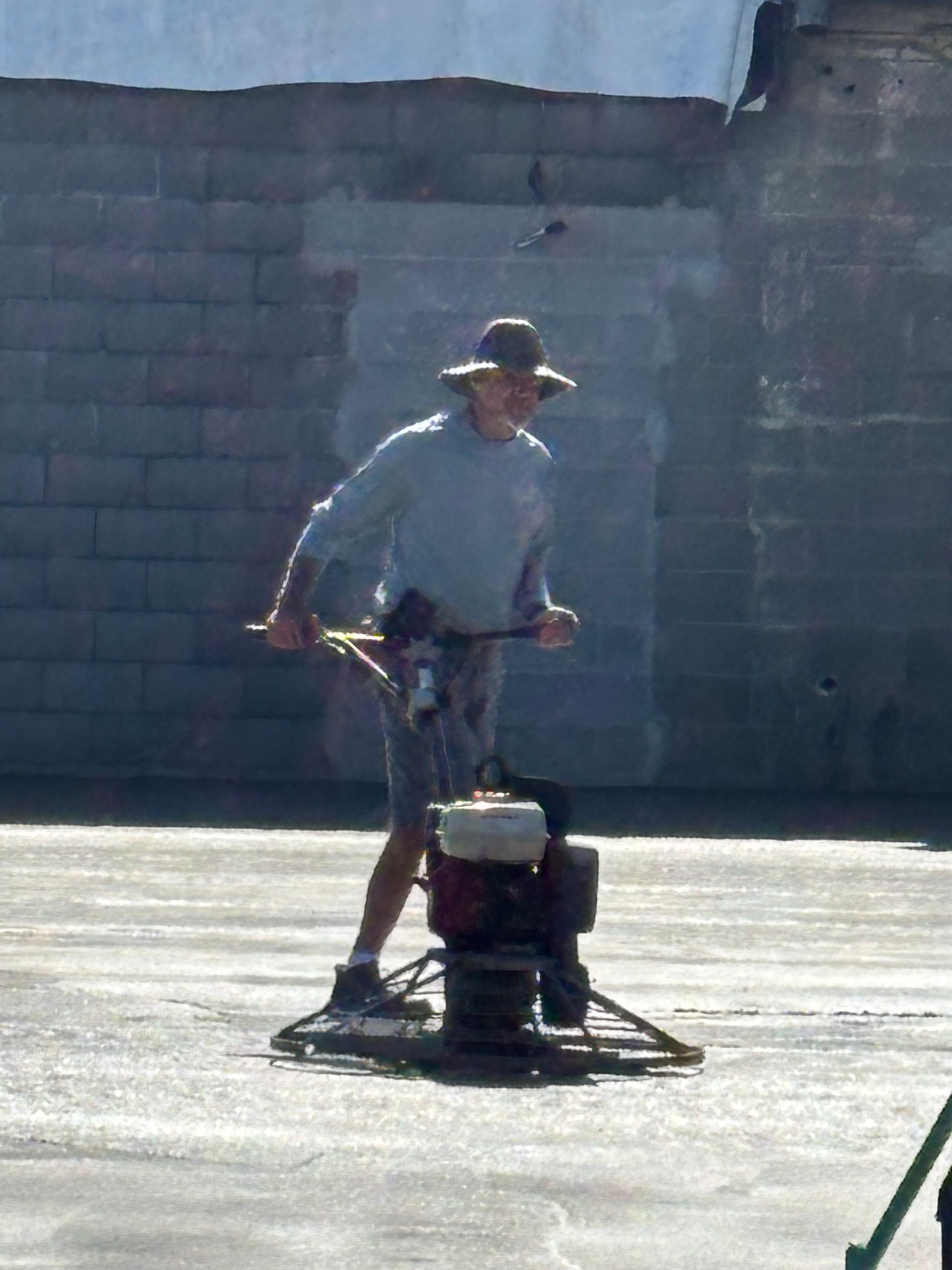 Person operating a walk-behind machine on a sunlit pavement, with a cinderblock wall in the background