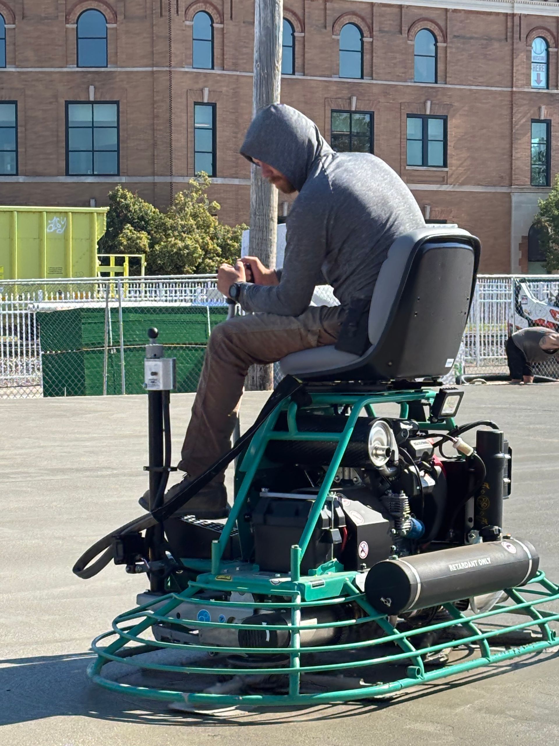 Person in a hoodie sits on a green tracked mobility device outdoors near a brick building.