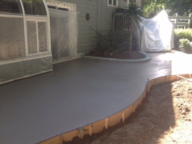 Freshly poured gray concrete patio curving around a house with a covered porch and white tarp-covered furniture