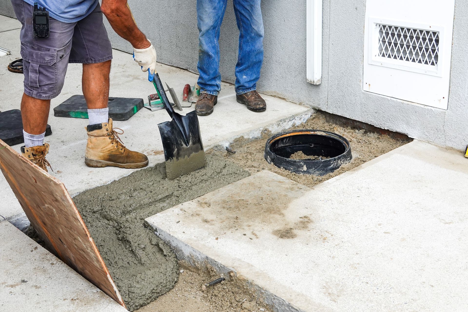 Workers pouring concrete around a round drain opening beside a building wall