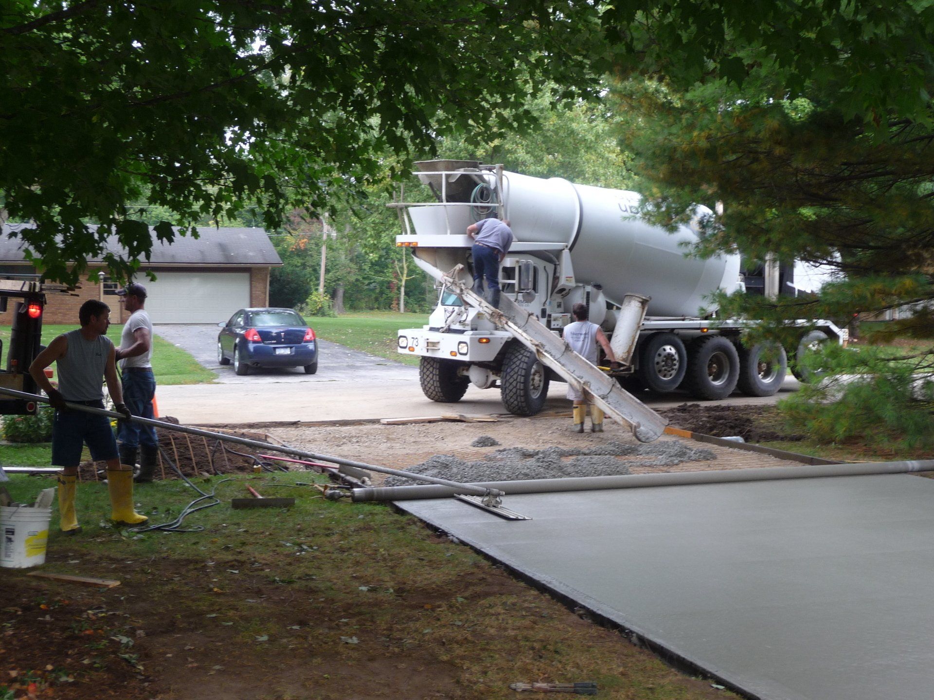 Workers pouring concrete from a truck onto a driveway near a house, with hoses and a wet slab in front.