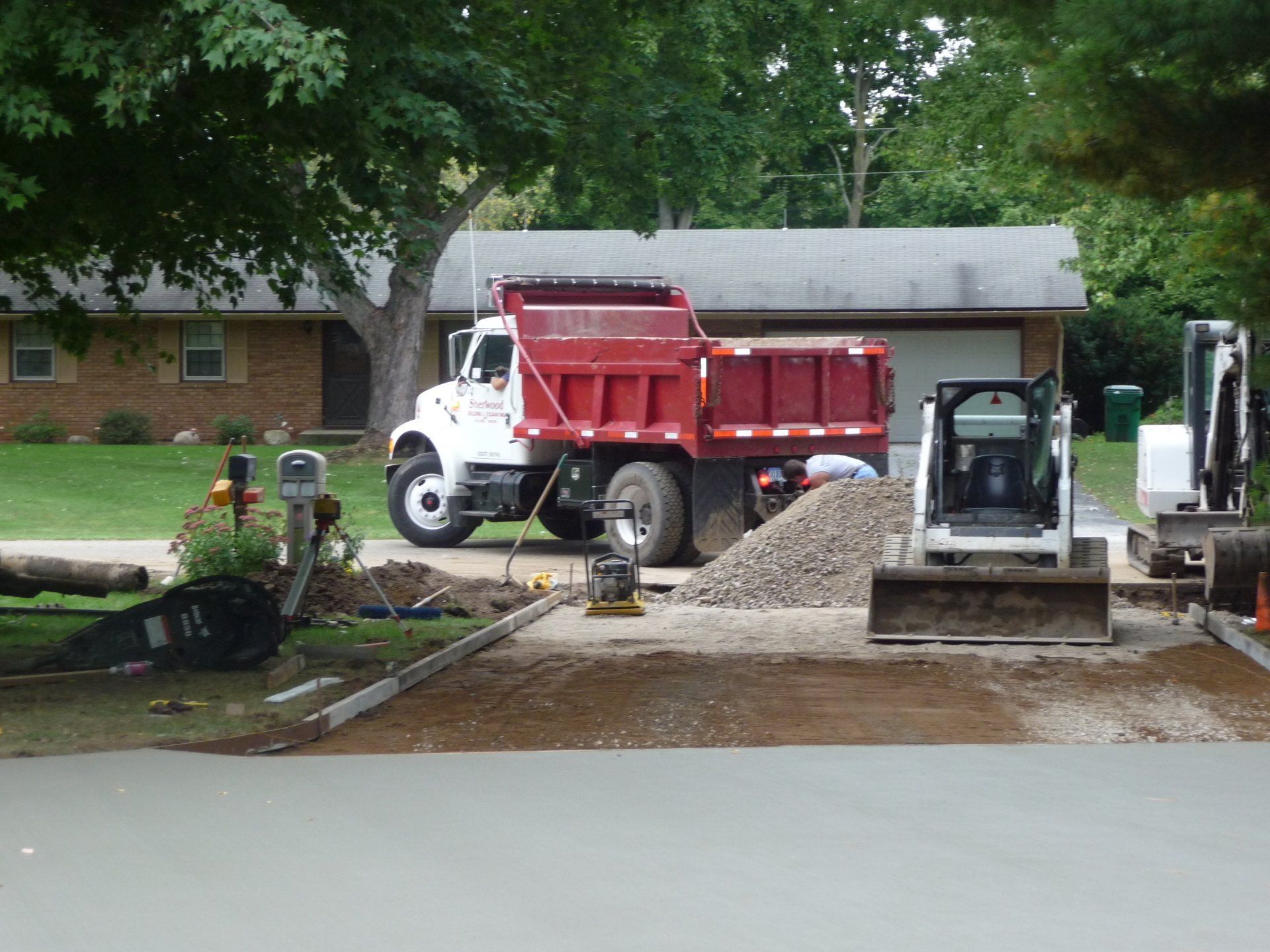 Construction vehicles grading a driveway with gravel beside a house and garage.