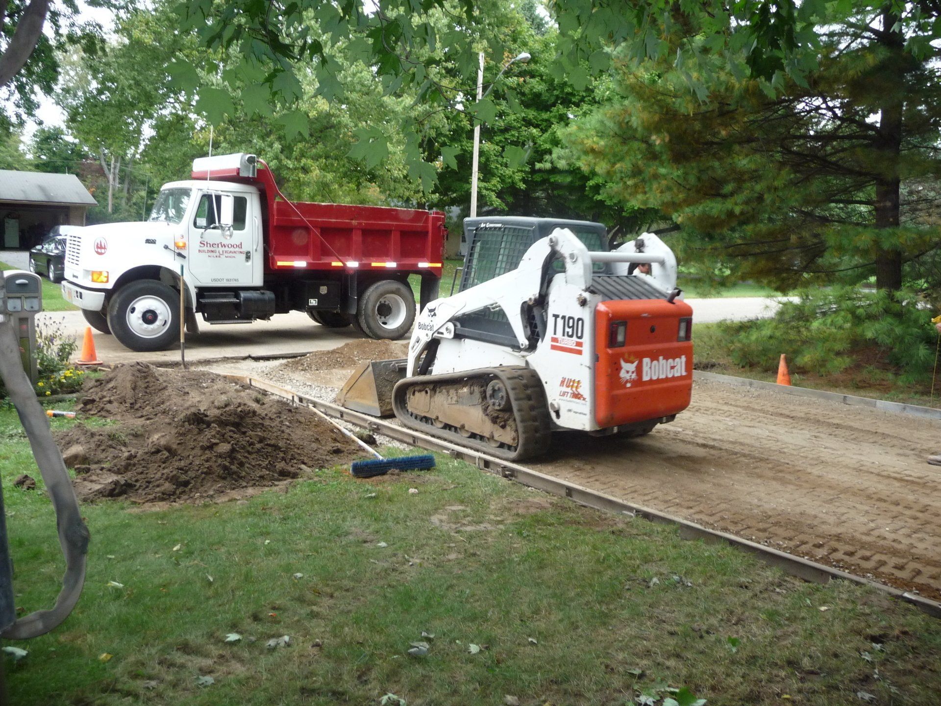Construction skid steer smoothing dirt beside a red dump truck on a tree-lined road