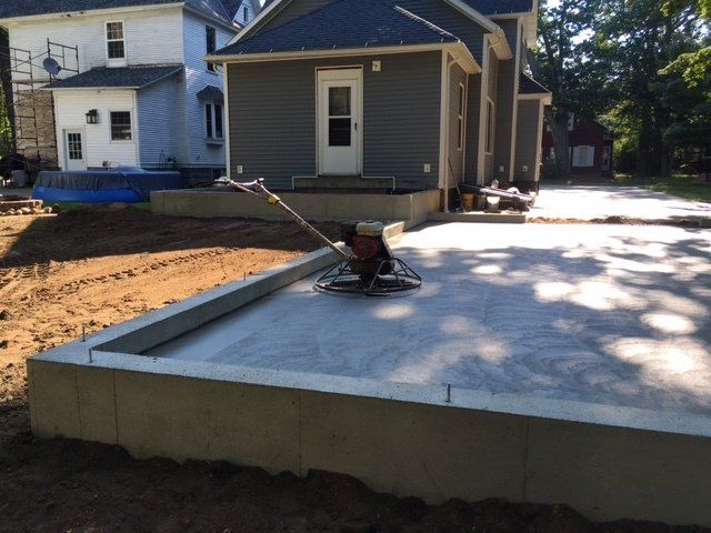 Concrete patio slab under construction beside a house, with a wheelbarrow and trowel on the fresh surface.
