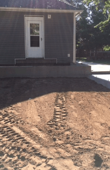 Front of a small gray house with a white door and a dirt driveway showing tire tracks.