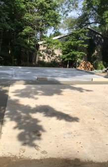 Concrete driveway under trees leading to a house, with sunlight and stacked firewood on the right.