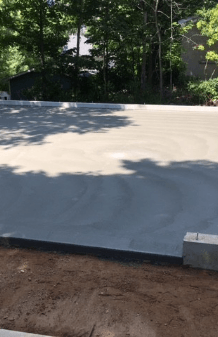 Freshly poured concrete foundation slab beside dirt and trees in a wooded outdoor area
