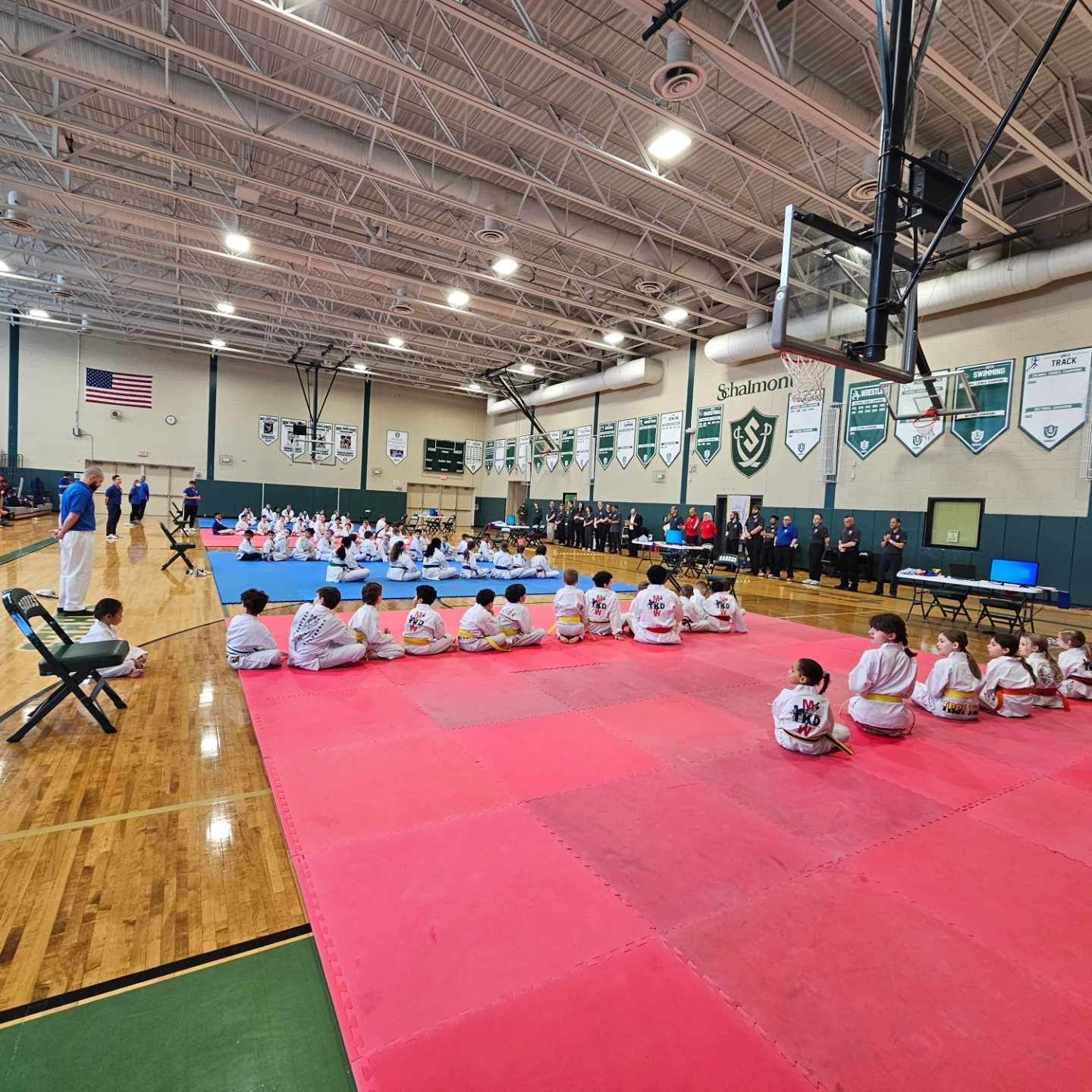 A group of children are sitting on a red mat in a gym.