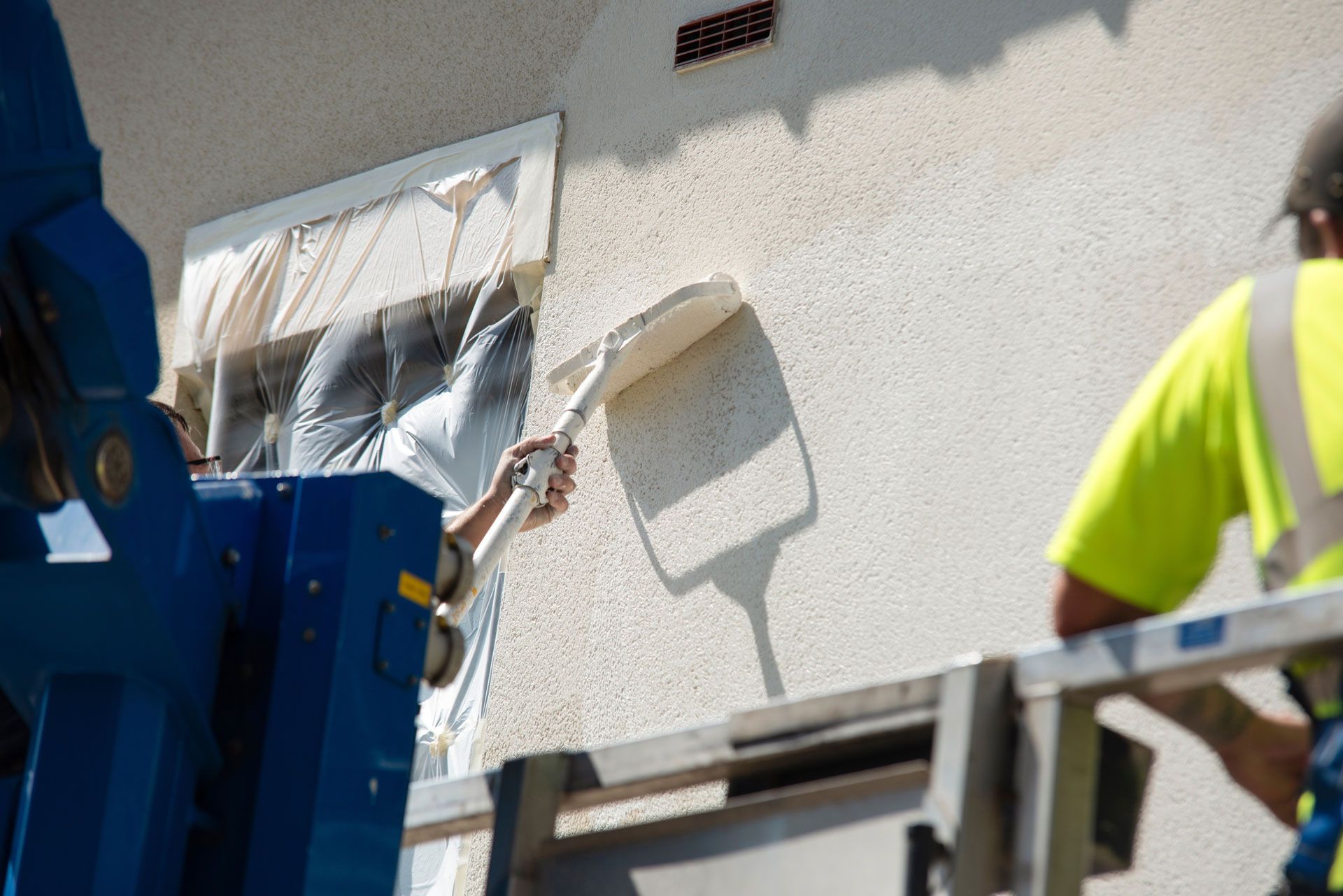 Workers plastering exterior wall near a window, using lift; natural light.