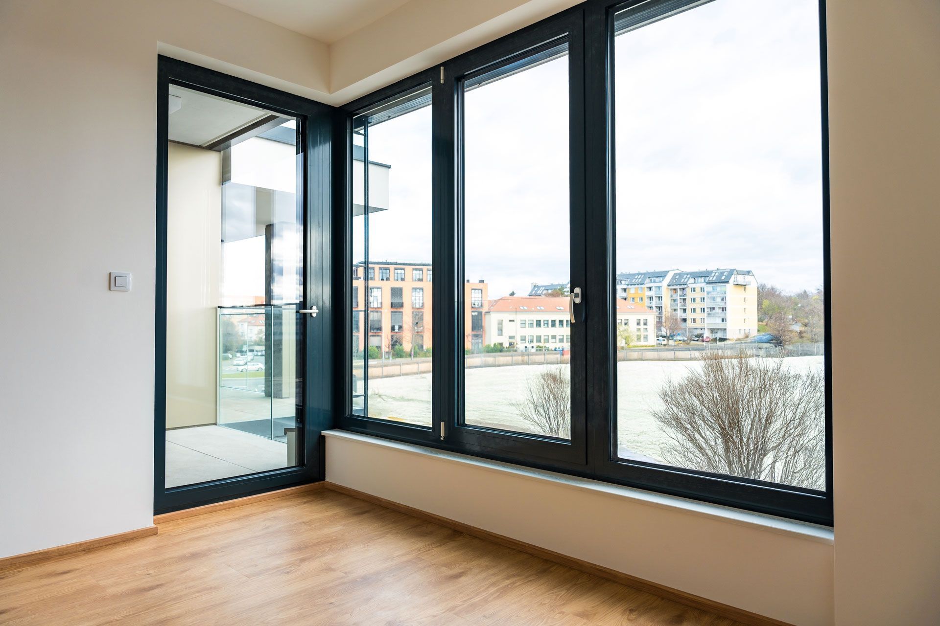 Empty room with large windows overlooking a city and a balcony. Hardwood floors and white walls.