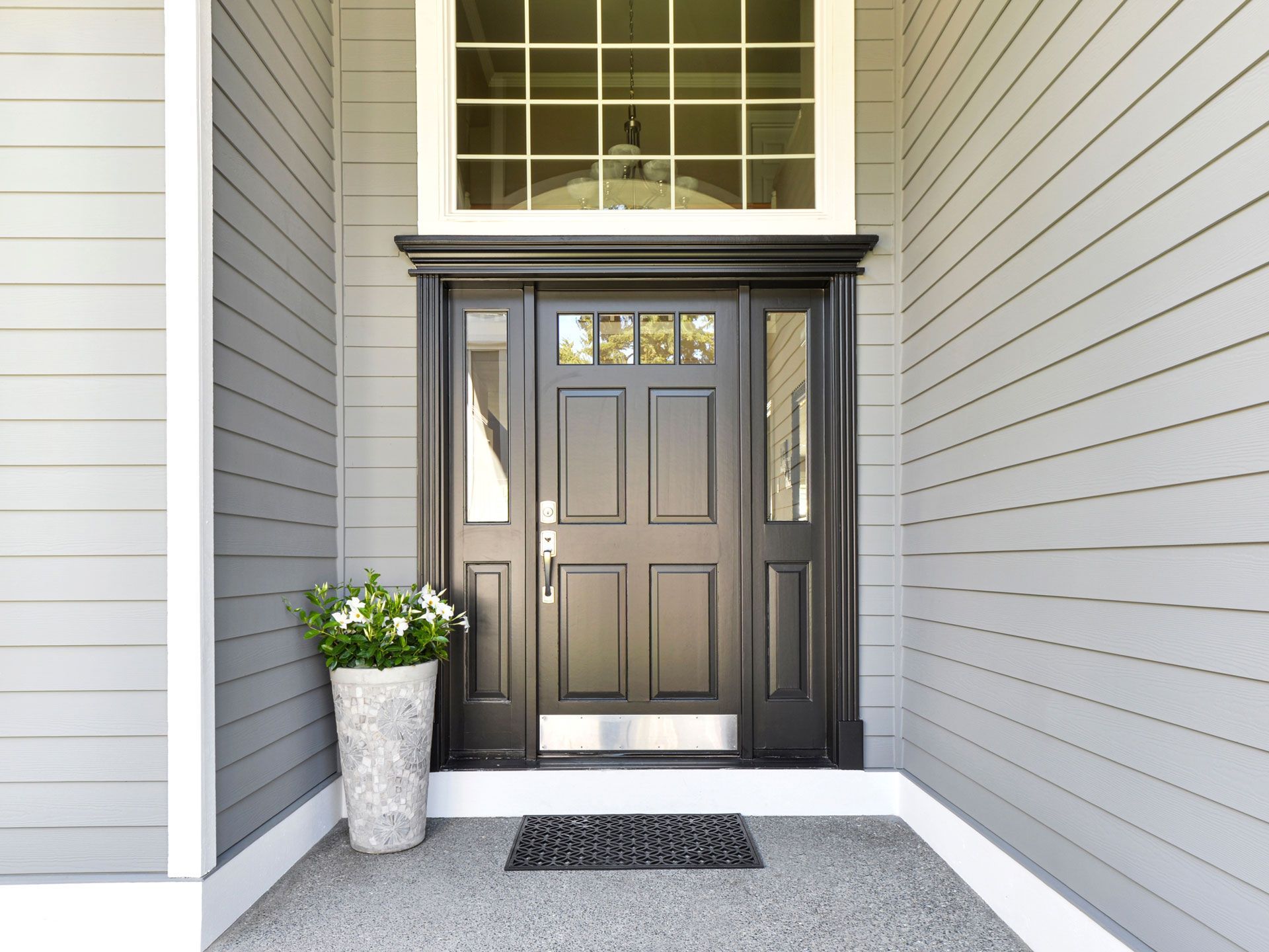 Black front door with sidelights and transom window, on a gray house with a potted plant.