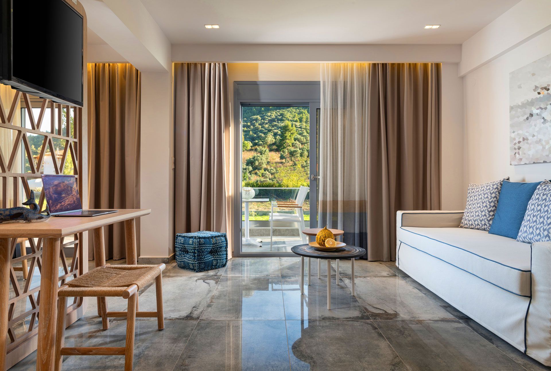 Living room with a desk, sofa, and a balcony view of trees. Gray tiled floor, beige and brown curtains.