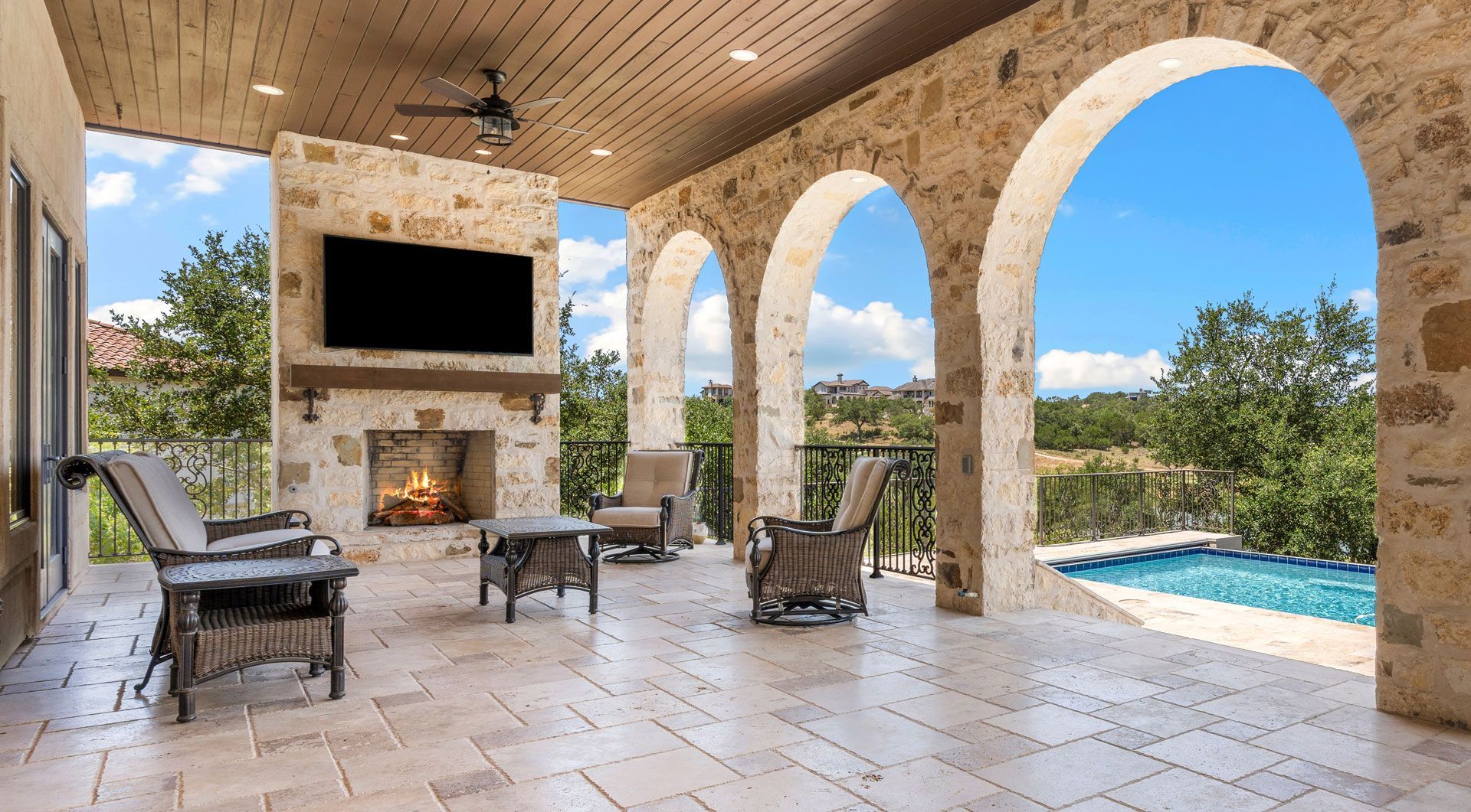 Covered outdoor patio with fireplace, TV, seating, and arches overlooking a pool and landscape.