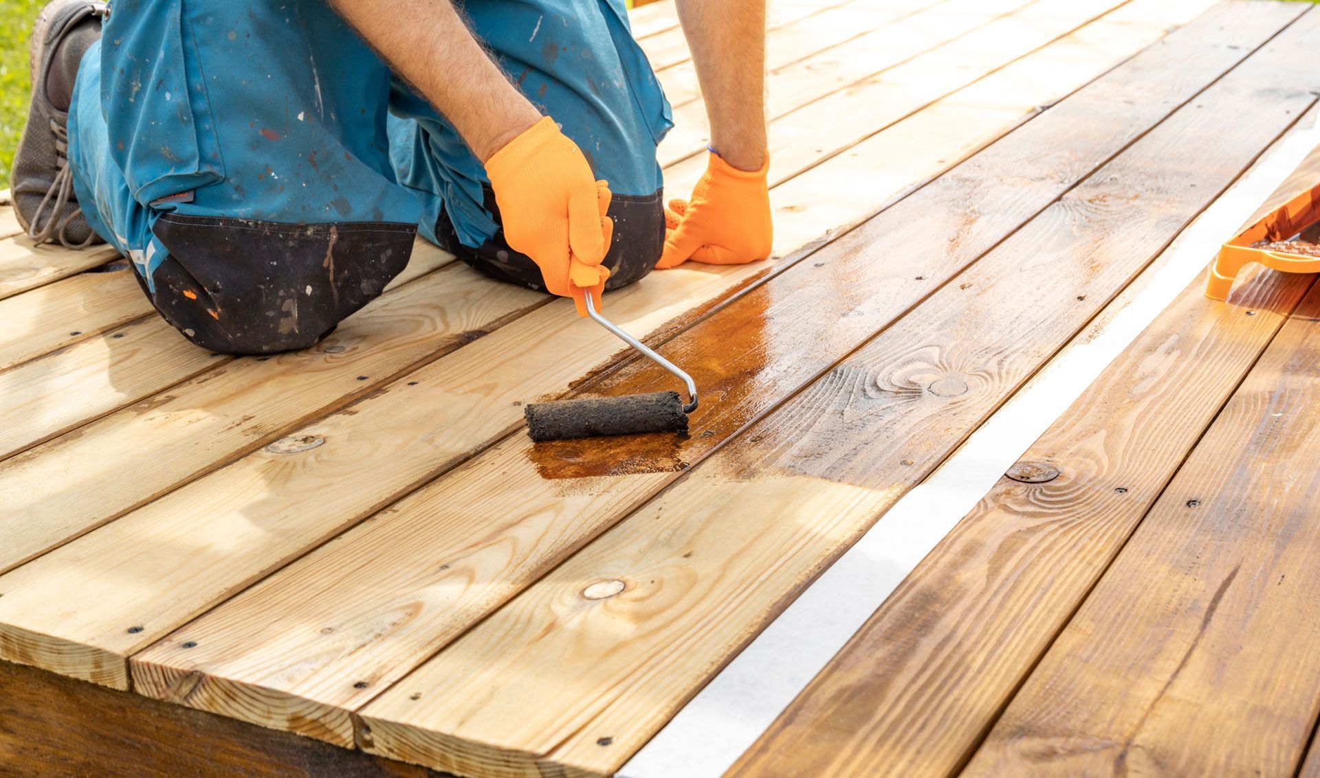Person staining a wooden deck with a roller, wearing gloves and blue overalls. Sunlight.