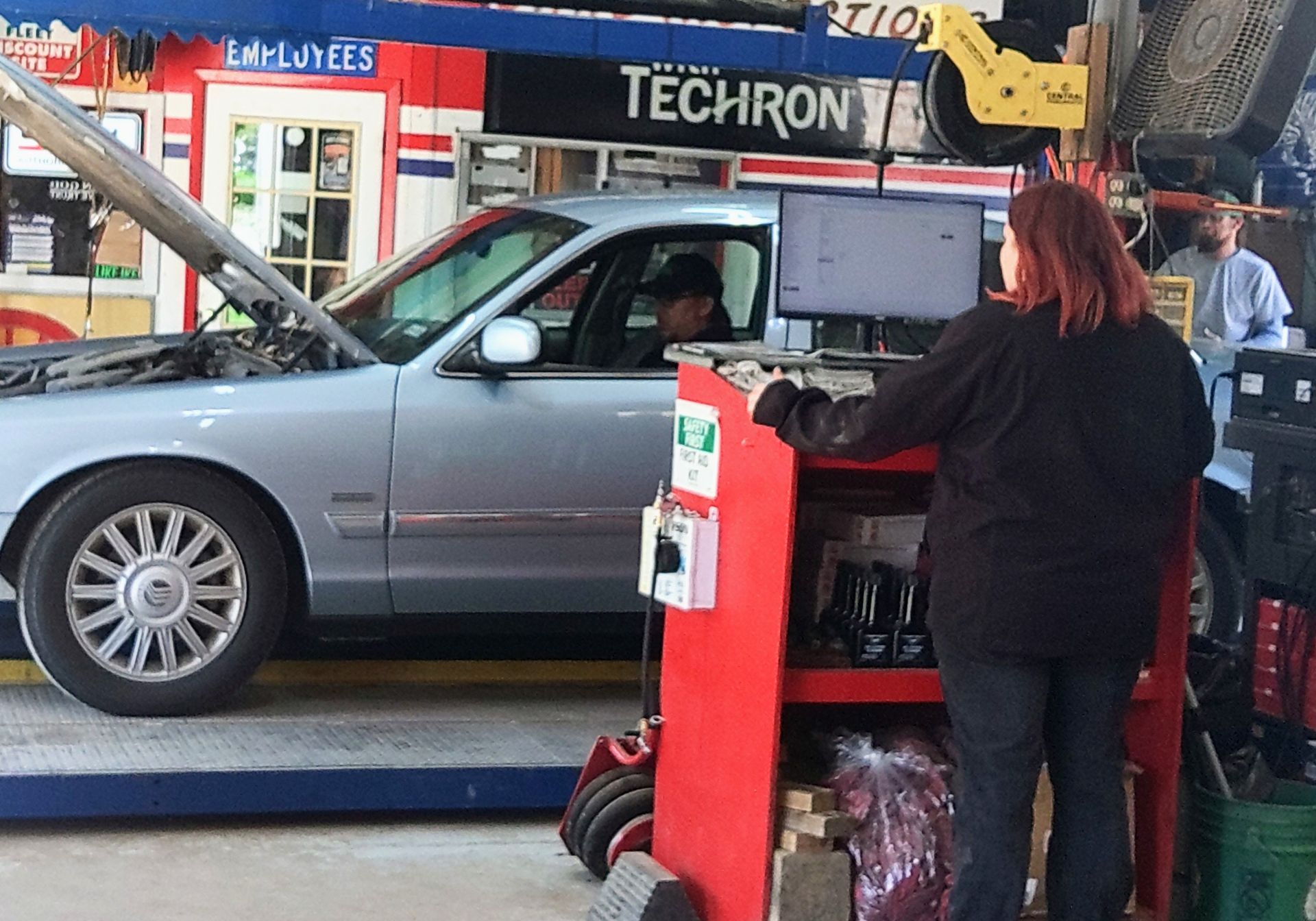 A woman is working on a car in front of a techron sign