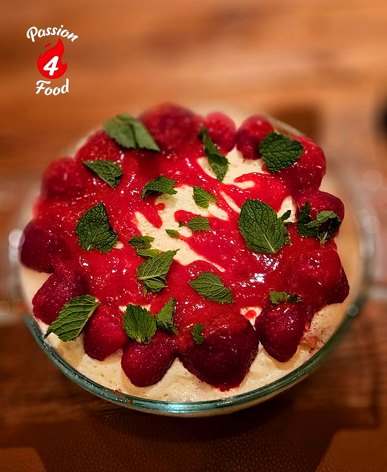Een dessert met aardbeien en muntblaadjes in een glazen kom, geserveerd op een houten tafel.