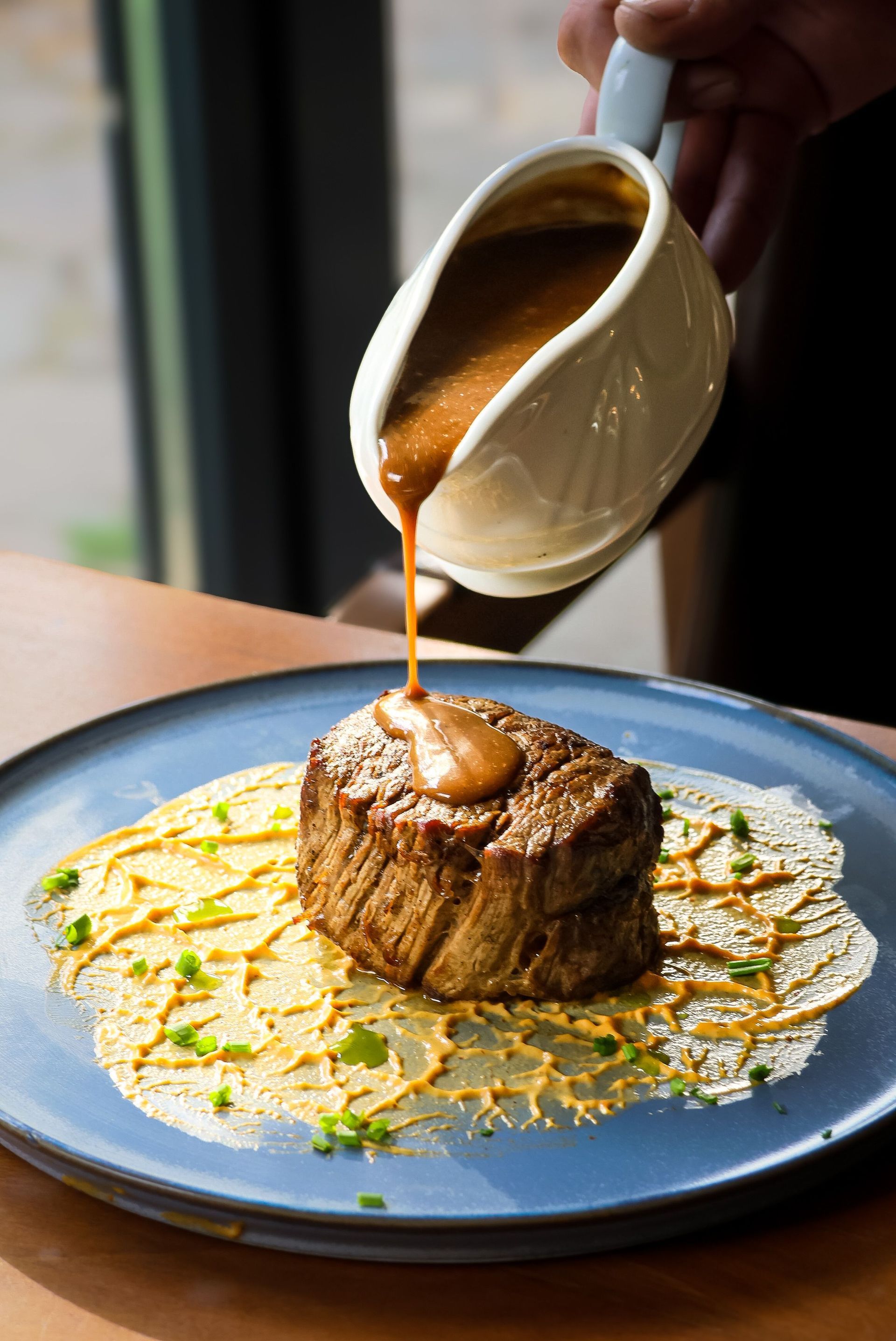 Sauce being poured over a seared steak on a blue plate, garnished with herbs and sauce drizzle