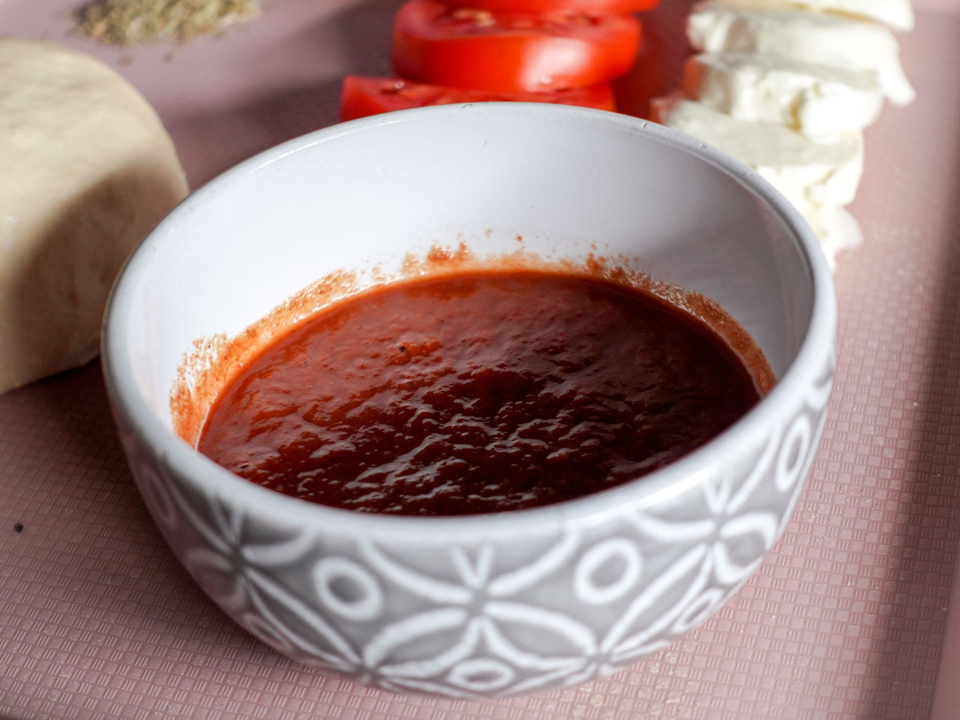 Een kom rode tomatensaus op een roze tafelkleed met gesneden groenten op de achtergrond.