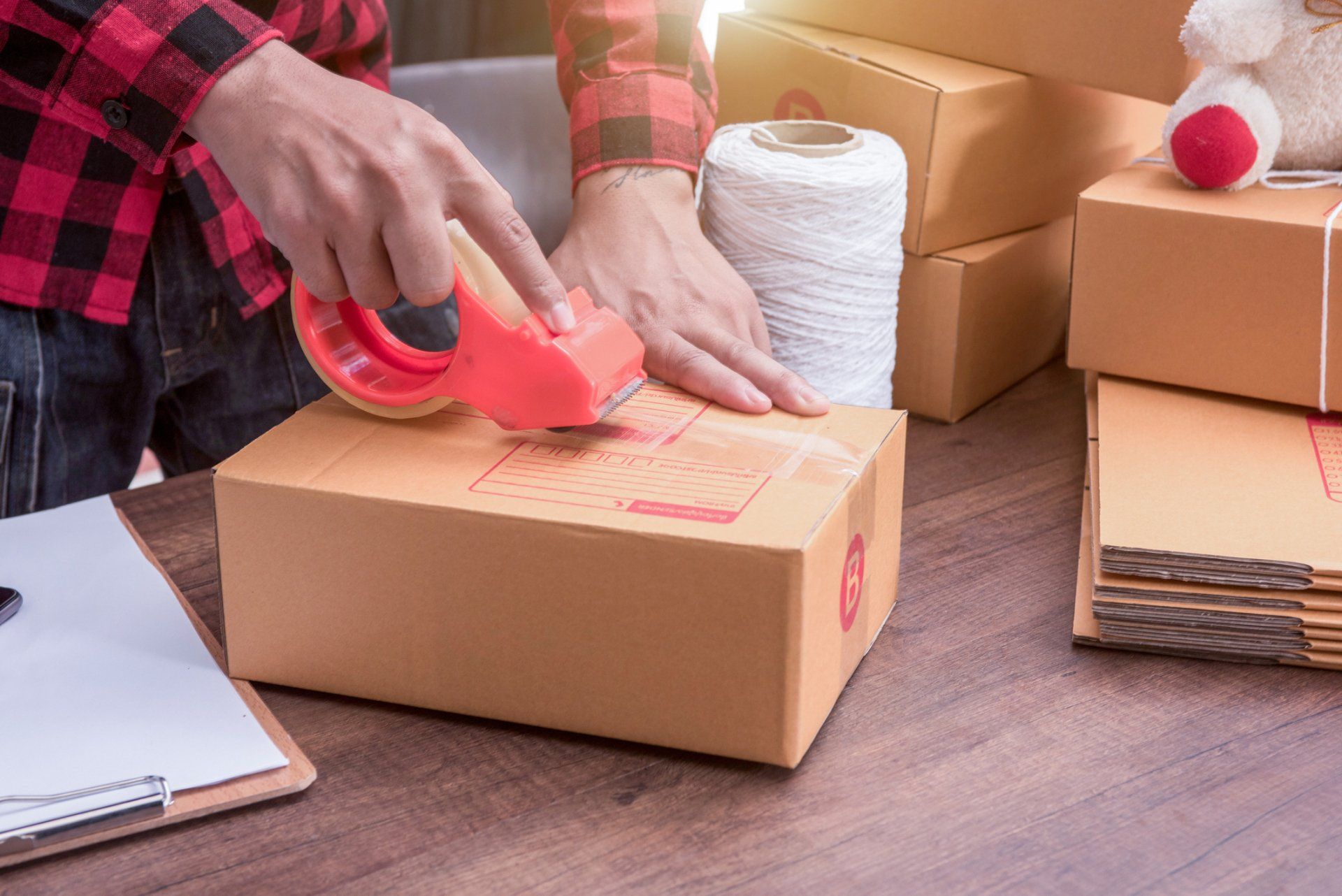 Person sealing a cardboard package with red tape at a table, with stacked boxes nearby