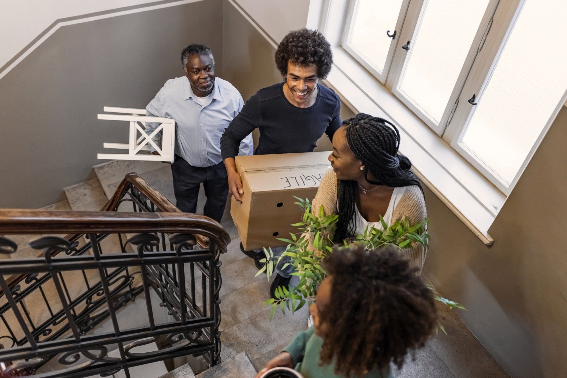 Three people carry a cardboard box up a stairwell beside a large window.