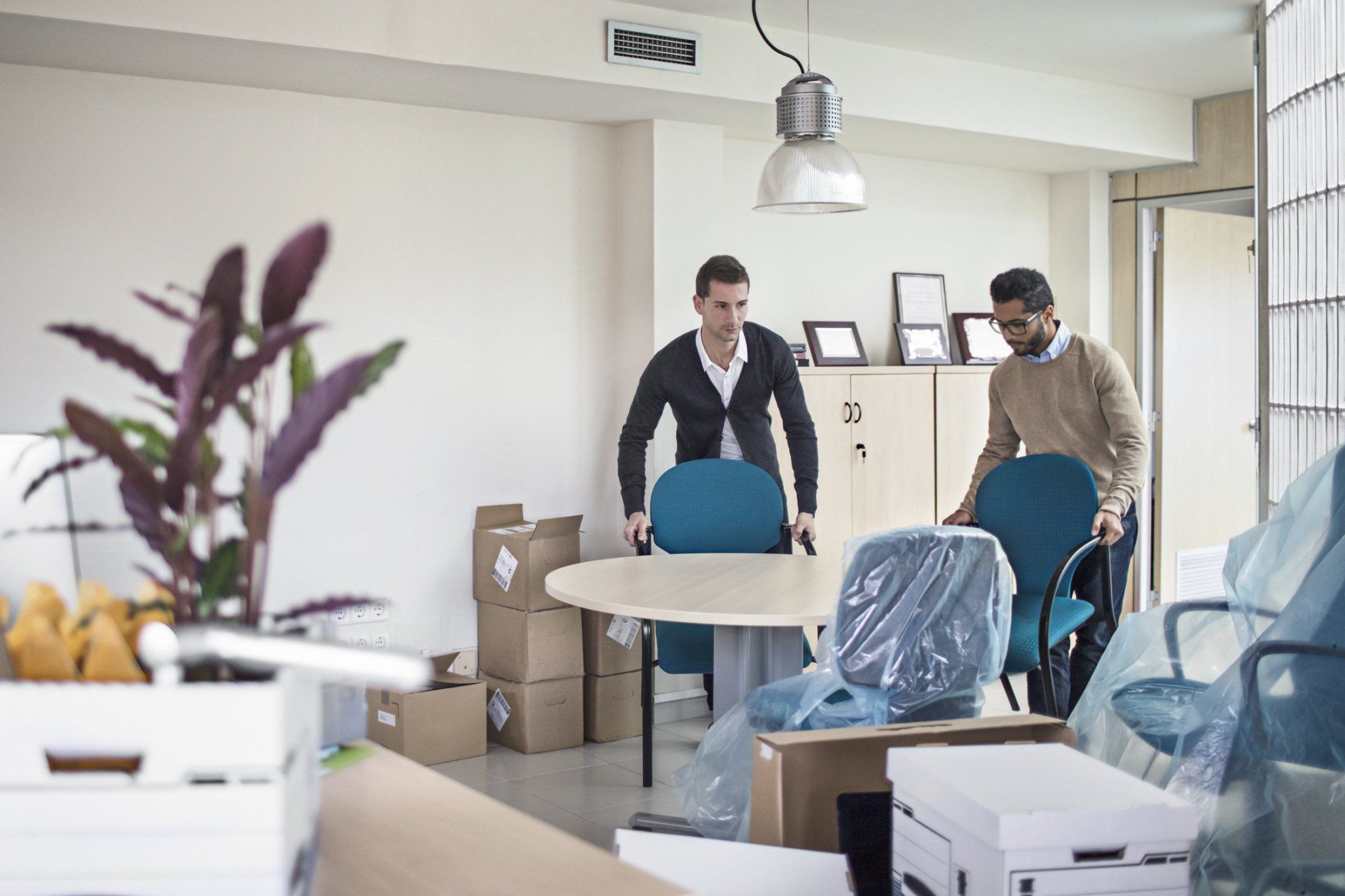 Two people moving office chairs in a bright room with boxes and covered equipment.