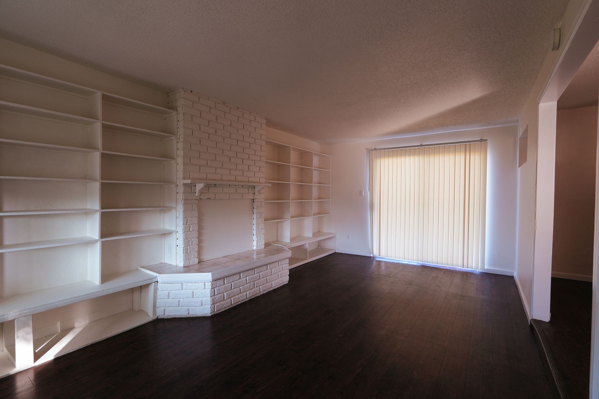 West Covina Empty living room with built-in white shelves, dark floor, and sheer curtains over sliding glass doors