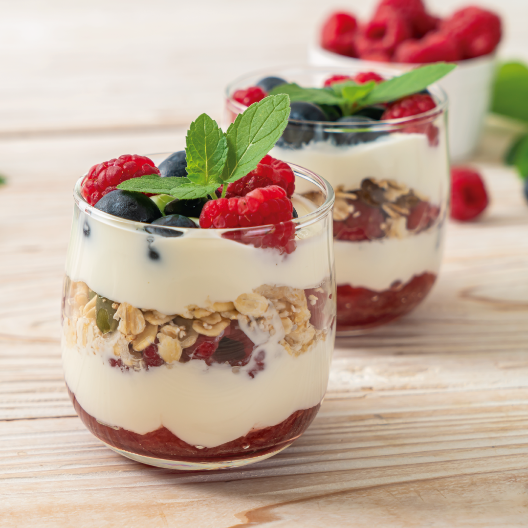 Dos vasos llenos de yogur, granola y frambuesas sobre una mesa de madera.