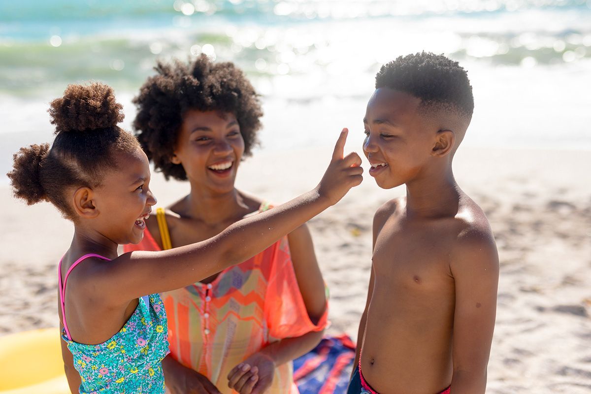 Three children on a sunny beach, smiling and playing near the water, one reaching toward another's face