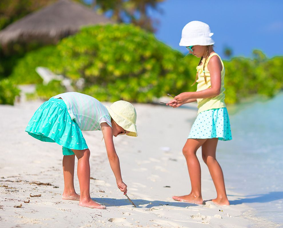 Two children in sun hats play on a sandy beach near turquoise water.