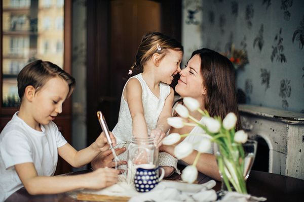 Child painting at a table while a woman kisses a smiling girl in a cozy kitchen