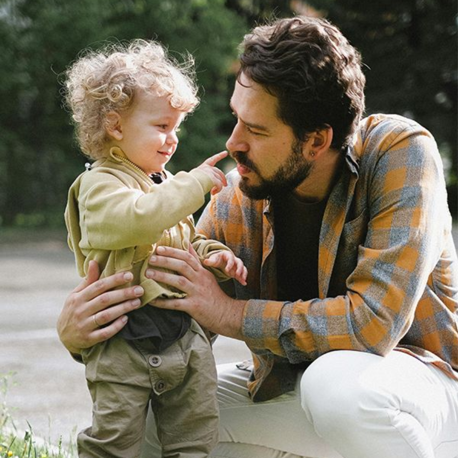 A man kneels outdoors with a smiling toddler, touching noses in a warm, candid moment.