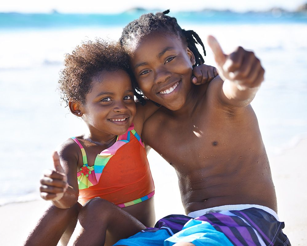 Two smiling children in swimwear sit on a sunny beach, one reaching toward the camera.