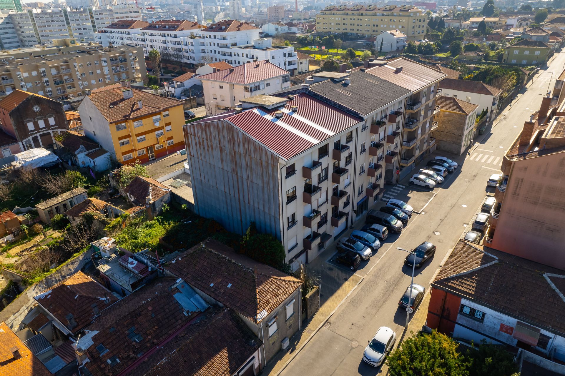 Vista aérea de edifícios ao longo de uma rua com carros estacionados e uma paisagem urbana ao fundo.